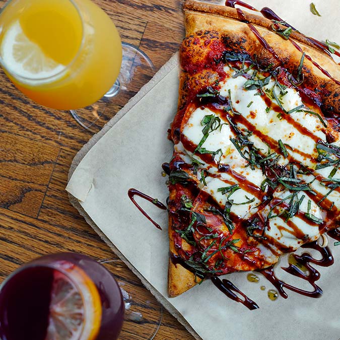 A slice of Pizza Beloit with cheese, basil, and sauce, paired with drinks at Downtown Den.