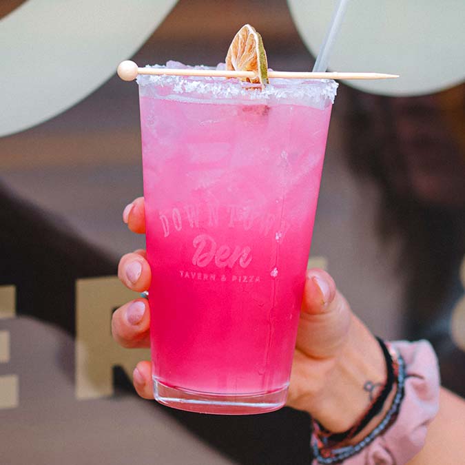 A hand holds a bright pink cocktail with ice at Downtown Den, rimmed with salt and dried lime.