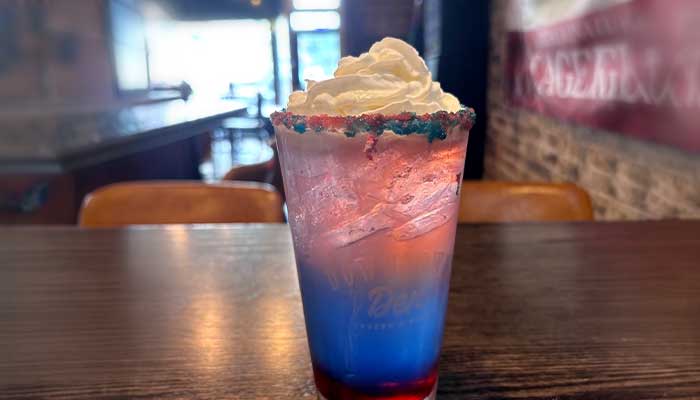 A layered red, white, and blue drink with whipped cream sits at Downtown Den bar in a cafe.