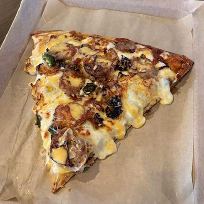 A slice of cheesy pizza with sausage and veggies served in a bar-style brown paper-lined box.
