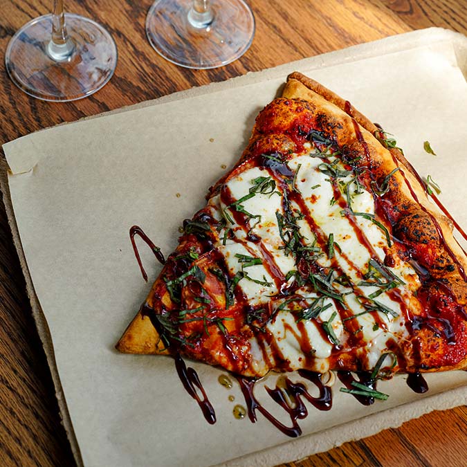 A slice of Pizza Beloit with mozzarella, basil, and balsamic glaze sits on parchment paper.